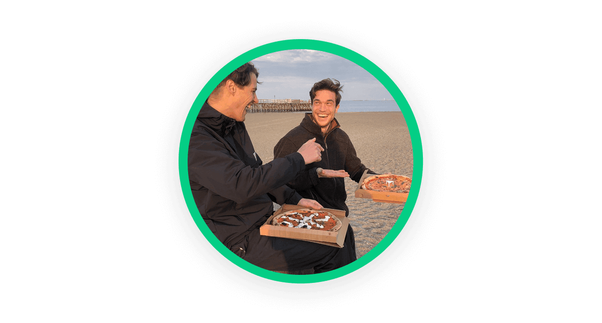A circular image showing a photo of two people at the beach, holding a pizza box in their hands and looking at each other and smiling